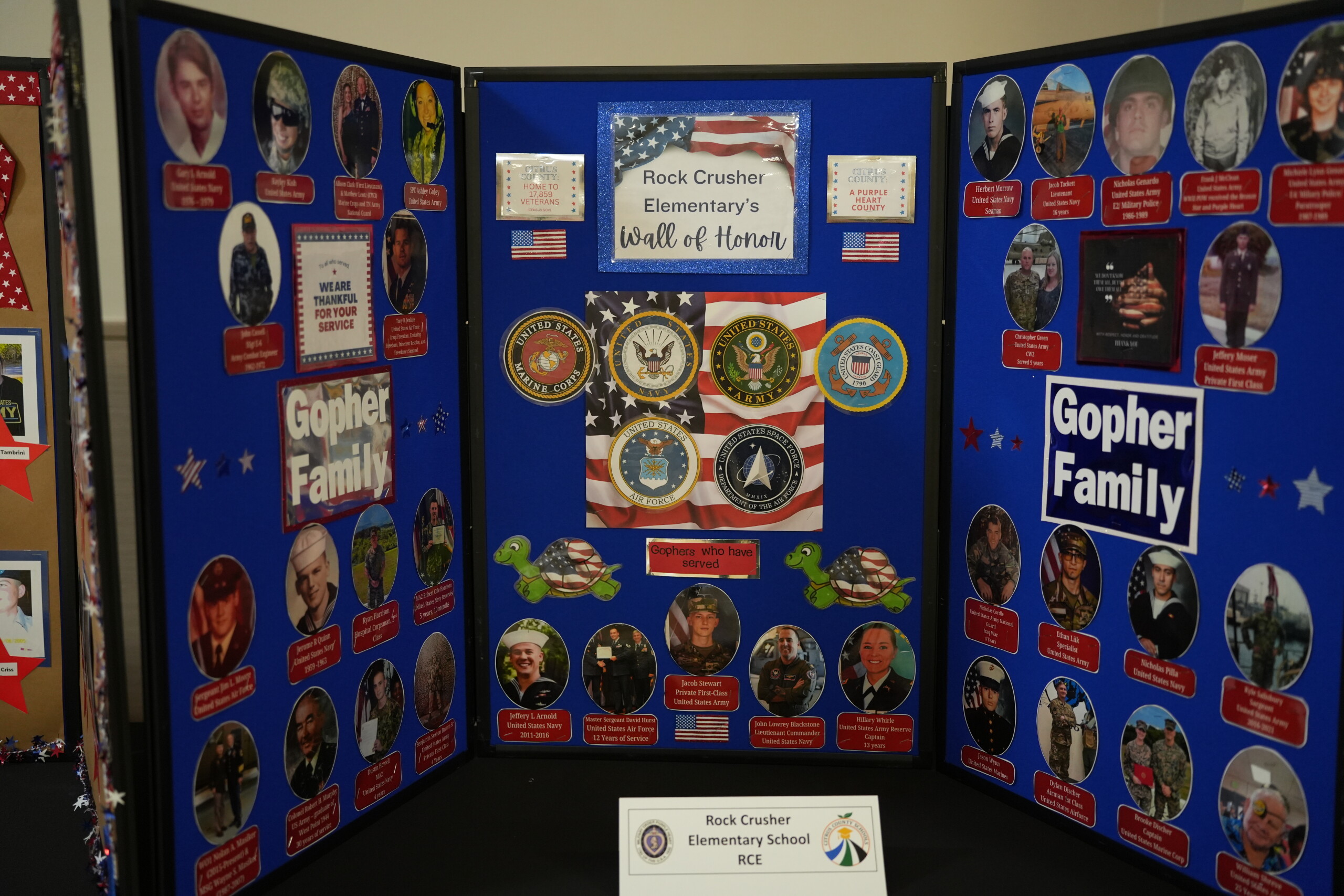 A trifold display board features Rock Crusher Elementary's Wall of Honor, decorated with photos of service members, military seals, and American flags.